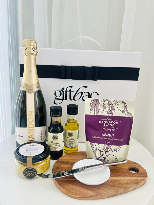 Gift bag with champagne, olives, and other items on a white surface | Luxury gift hampers Australia – beautifully curated gourmet hampers, wine gift boxes, and pamper hampers for all budgets. Perfect for birthdays, anniversaries, corporate gifts & special occasions. Gift Bae offers Perth same-day delivery & Australia-wide shipping. Shop the best gift hampers online today!